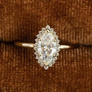 Könnte beinhalten: Ein goldener Ring mit einem Marquise-Diamanten, der in einem Halo aus kleineren Diamanten gefasst ist. Der Ring befindet sich in einer braunen Samt-Ringbox.