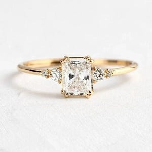 Könnte beinhalten: Ein goldener Ring mit einem rechteckigen, klaren Edelstein als Mittelpunkt. Zwei kleinere, dreieckige Edelsteine flankieren den Hauptstein. Das Band ist dünn und goldfarben und schafft ein zartes und elegantes Design.