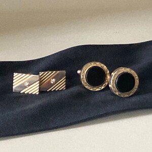 May include: Two pairs of cufflinks on a dark blue surface. One pair is rectangular with a silver and gold design and a small diamond. The other pair is round with a black center and a gold textured border.