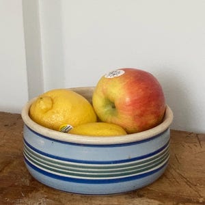 May include: A ceramic bowl with a blue and green striped design holds a red apple, a lemon, and a smaller yellow fruit. The bowl sits on a wooden surface, with a white wall in the background.