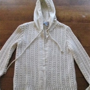 May include: A cream-coloured hooded cardigan with a drawstring closure. The cardigan features a textured knit pattern and long sleeves. The hood and front panel are a solid colour, while the body has a vertical open-knit design.