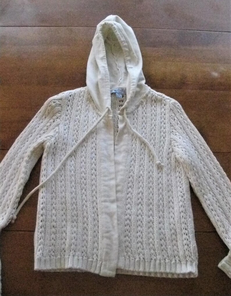 May include: A cream-coloured hooded cardigan with a drawstring closure. The cardigan features a textured knit pattern and long sleeves. The hood and front panel are a solid colour, while the body has a vertical open-knit design.