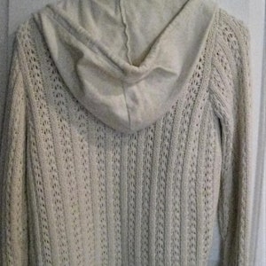 May include: Cream-coloured hooded sweater with a textured knit design. The back of the sweater is visible, showing the hood and the intricate openwork pattern. The sweater is hanging on a white hanger.
