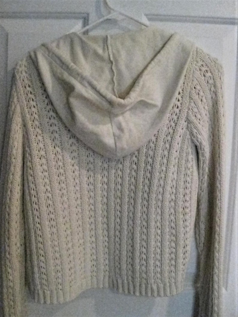 May include: Cream-coloured hooded sweater with a textured knit design. The back of the sweater is visible, showing the hood and the intricate openwork pattern. The sweater is hanging on a white hanger.