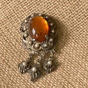 May include: A silver brooch with an amber-colored glass stone and dangling pearl accents.