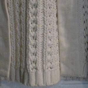 May include: Close-up of a cream-coloured knitted sweater with a cable knit design. The sweater features a textured pattern and a smooth, solid panel on the side. The bottom hem is ribbed.