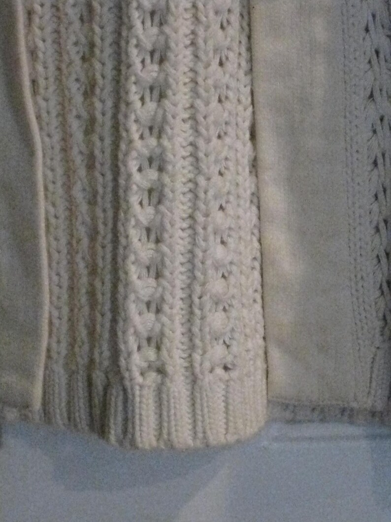 May include: Close-up of a cream-coloured knitted sweater with a cable knit design. The sweater features a textured pattern and a smooth, solid panel on the side. The bottom hem is ribbed.