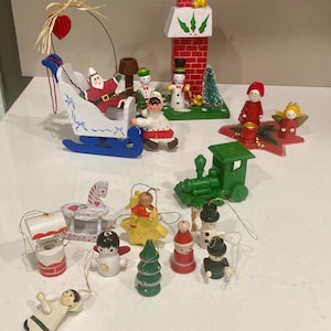 May include: A collection of Christmas ornaments, including a Santa Claus in a sleigh, a red brick chimney, a green train, angels, snowmen, and other festive figurines. The ornaments are made of wood and painted in bright colors.