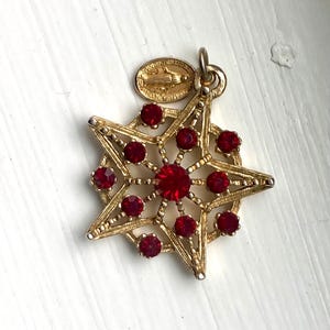 May include: Gold-tone pendant featuring a star-shaped design with red rhinestone accents. The pendant has a smaller oval charm with a religious image.