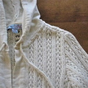 May include: Cream-coloured knitted hoodie with a drawstring and hood. The sweater features a textured, open-weave design and a label that reads "bdg". The hoodie is a neutral colour and appears to be made of a soft material.