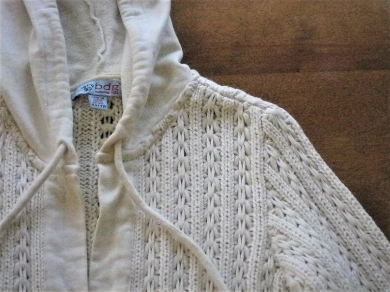 May include: Cream-coloured knitted hoodie with a drawstring and hood. The sweater features a textured, open-weave design and a label that reads "bdg". The hoodie is a neutral colour and appears to be made of a soft material.