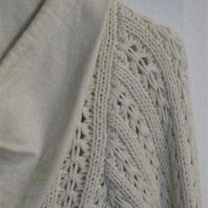 May include: Detail of a cream-coloured knitted sweater. The sleeve displays an intricate openwork design, contrasting with the smooth, solid collar. The image highlights the texture and craftsmanship of the knitwear, showcasing the garment's detail.