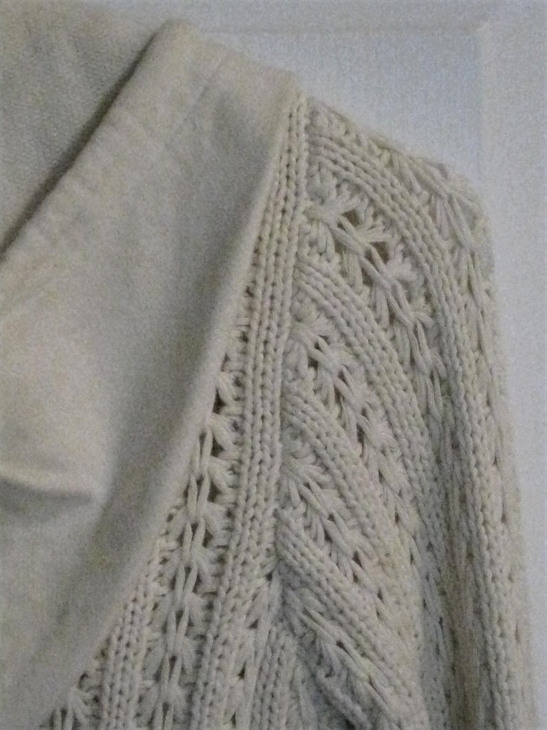 May include: Detail of a cream-coloured knitted sweater. The sleeve displays an intricate openwork design, contrasting with the smooth, solid collar. The image highlights the texture and craftsmanship of the knitwear, showcasing the garment's detail.