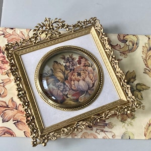 May include: A gold-colored, ornate picture frame with a floral print. The frame has a square shape with a circular inset containing a floral painting. The frame is decorated with leaf and vine details.