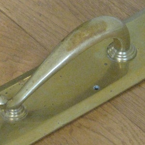 May include: A brass door handle with a curved, arched design. The handle is mounted on a rectangular plate. The hardware has a polished finish and is set against a wooden background.