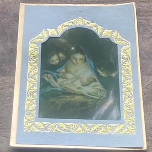 May include: A religious card featuring a depiction of the Virgin Mary holding baby Jesus, surrounded by angels. The image is framed by a light blue border with a gold filigree design. The card is rectangular and appears to be made of paper.