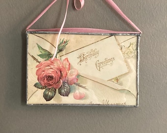 May include: Vintage framed artwork featuring a birthday greeting card with a pink rose illustration. The card is inside an envelope with the words "Birthday Greetings" and is suspended by a pink ribbon.