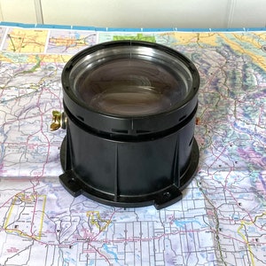 May include: A large, black lens with a clear glass element. The lens is cylindrical with a metal adjustment screw and sits on a detailed map. The lens is likely for a camera or other optical device.