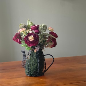 May include: A bouquet of burgundy and cream colored artificial flowers in a black wire teapot vase. The vase is filled with green moss.
