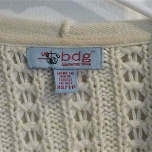May include: Close-up of a cream-coloured knit sweater with a textured pattern. A blue tag reads "bdg GUARANTEED TOUGH" and "MADE IN INDIA". The sweater is XS/TP size.