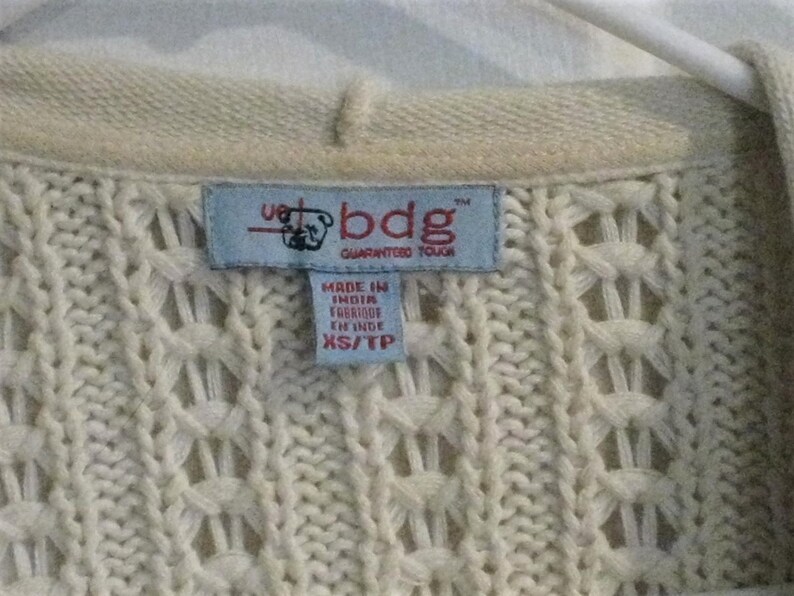 May include: Close-up of a cream-coloured knit sweater with a textured pattern. A blue tag reads "bdg GUARANTEED TOUGH" and "MADE IN INDIA". The sweater is XS/TP size.