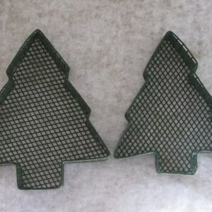 May include: Two dark green, Christmas tree-shaped metal mesh forms. The forms have a diamond-shaped mesh pattern within a solid outline. The forms are placed on a light-colored, textured surface.