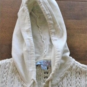 May include: Cream-coloured hooded sweater with a textured knit pattern. The hood is made of a softer material and has a drawstring. The label reads "bdg" and "gathered york". The sweater is a size XS/TP.