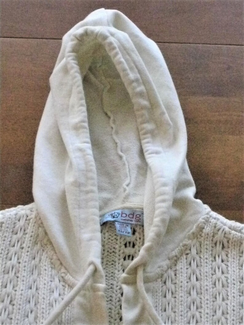 May include: Cream-coloured hooded sweater with a textured knit pattern. The hood is made of a softer material and has a drawstring. The label reads "bdg" and "gathered york". The sweater is a size XS/TP.