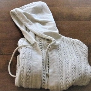 May include: A cream-coloured hooded cardigan. The sweater has an open-knit texture, a drawstring hood, and a button-up front. It is folded on a wooden surface.