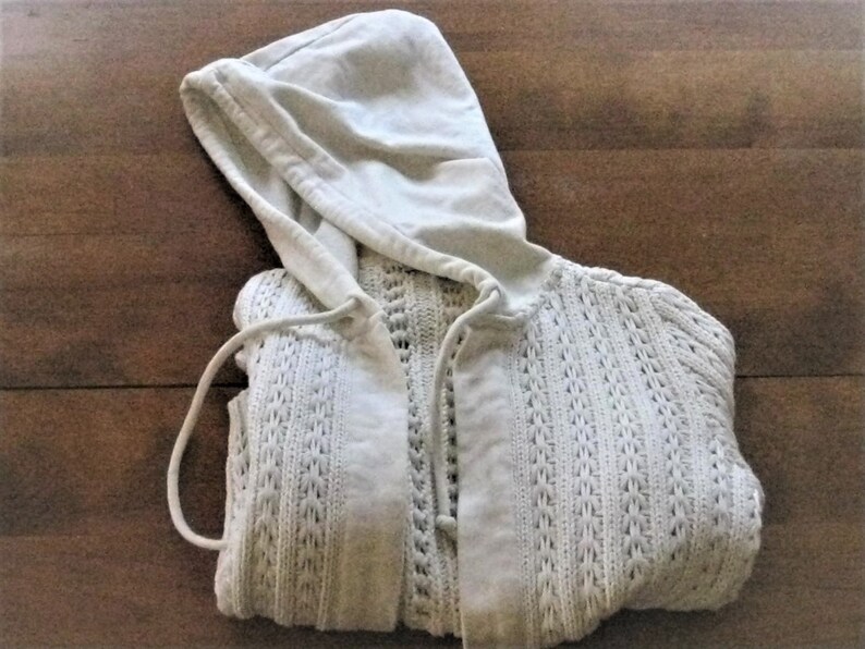 May include: A cream-coloured hooded cardigan. The sweater has an open-knit texture, a drawstring hood, and a button-up front. It is folded on a wooden surface.