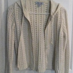 May include: Cream-coloured, open-front hooded sweater with a textured knit design. The sweater features long sleeves, a drawstring hood, and a tag that reads "bdg". The garment is hanging on a white hanger against a white door.