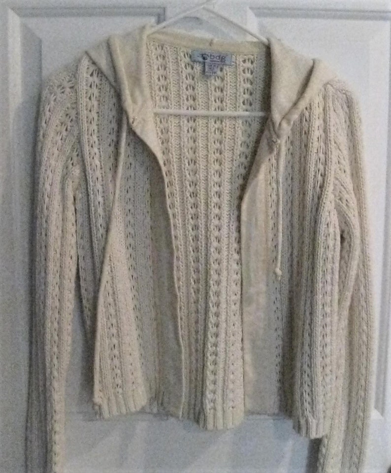 May include: Cream-coloured, open-front hooded sweater with a textured knit design. The sweater features long sleeves, a drawstring hood, and a tag that reads "bdg". The garment is hanging on a white hanger against a white door.