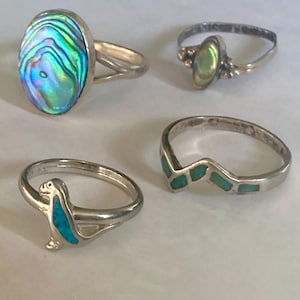 May include: Four silver rings. One ring features an oval abalone shell. Another has a small abalone shell. A third ring has a turquoise bird design. The fourth ring has a zig-zag pattern with turquoise inlays. Jewelry.