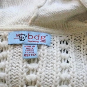 May include: Close-up of a cream-coloured knit sweater with a hood. The sweater features a textured pattern and a blue label with the text "bdg GUARANTEED TOUGH" and "MADE IN INDIA FABRIQUE EN INDE XS/TP".