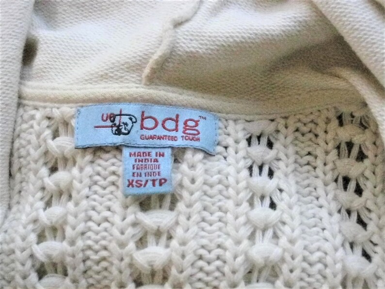 May include: Close-up of a cream-coloured knit sweater with a hood. The sweater features a textured pattern and a blue label with the text "bdg GUARANTEED TOUGH" and "MADE IN INDIA FABRIQUE EN INDE XS/TP".