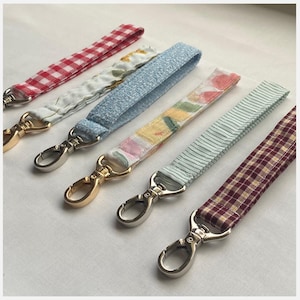 Puede incluir: Seis cordones de muñeca de tela con varios patrones, incluyendo cuadros rojos, florales, azules texturizados y diseños a rayas. Cada cordón tiene un cierre giratorio plateado y un cierre giratorio dorado.