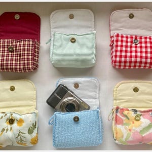 May include: A selection of small fabric pouches with magnetic closures. The pouches display diverse patterns, such as gingham, stripes, floral, and solid colours. One pouch contains a Canon camera. The pouches are open, showing their interior lining.