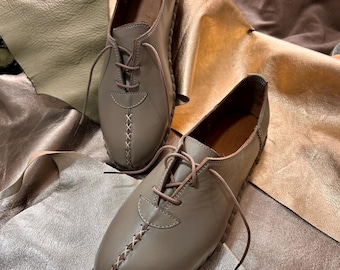 Op de afbeelding: Een paar grijze leren veterschoenen met een gestikt detail rond de neus en zool. De schoenen liggen op een lichtbruin lederen oppervlak.