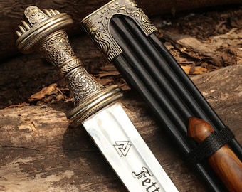 May include: A detailed close-up of a decorative sword with an ornate, gold-colored handle and a silver blade. The blade has etched text and a symbol. The sword rests next to a black sheath with decorative gold accents.