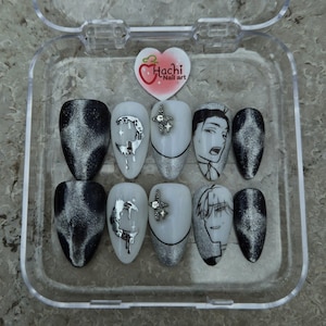 May include: A set of ten artificial nails in a clear plastic case. The nails feature various designs, including black and silver cat-eye effect, silver moons, cross-shaped gems, and anime-style character art. The case has a heart-shaped logo that says "Hachi Nail Art".