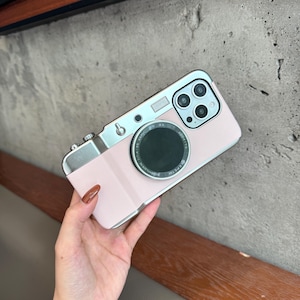 以下が含まれることがあります： ヴィンテージカメラに似せてデザインされたピンク色の携帯電話ケース。ケースは、シルバーカラーのトップとレンズディテール、大きな黒い円形のレンズを備えています。携帯電話ケースには、3つのレンズカメラシステムがあります。