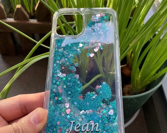 May include: A clear phone case with a glittery liquid inside. The glitter is blue and pink, and there are small hearts and stars mixed in. The name "Jean" is written on the case.