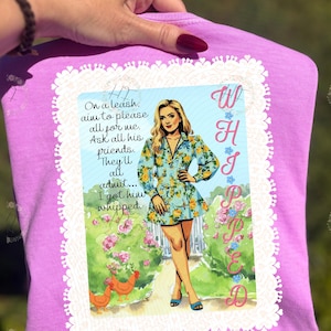 Puede incluir: Una camiseta de color lila con un gráfico de una mujer con un vestido floral, texto y un borde decorativo. El texto incluye las palabras "Whipped" y "On a leash, aim to please all for me."