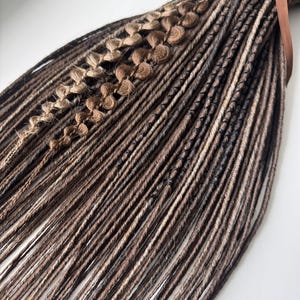 Puede incluir: Un conjunto de rastas marrones y negras con detalles en marrón claro y secciones decorativas. Las rastas están atadas con una cinta marrón. Las extensiones de cabello son una mezcla de tonos oscuros y claros.