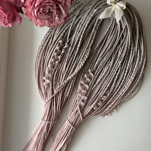 May include: Two bundles of dreadlocks in shades of gray and pink, tied together with string. The dreadlocks are decorated with white stitching and small embellishments. A white bow is attached to the top. The background includes pink roses.