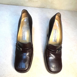 May include: A pair of brown leather heeled shoes with a square toe and a small buckle detail. The shoes have the "Dolce & Gabbana" label inside. The heels are approximately 2 inches high.