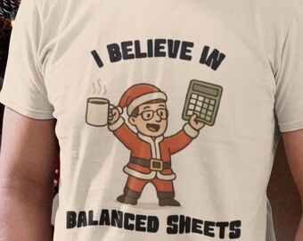 May include: Beige t-shirt with the text "I Believe in Balanced Sheets" and a cartoon Santa Claus holding a coffee mug and calculator. The Santa illustration is in red and white. The person is wearing blue jeans.