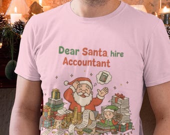 May include: A light pink t-shirt with the text "Dear Santa, hire Accountant" above a graphic of Santa Claus and an elf surrounded by gifts and paperwork. The shirt is a festive holiday design.