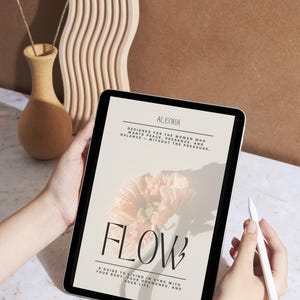 May include: A digital tablet displaying the cover of a book titled "FLOW" with a floral illustration. The text reads "ALEORA" and "A guide to living in sync with your body, your hormones, and your life." A stylus is held nearby.