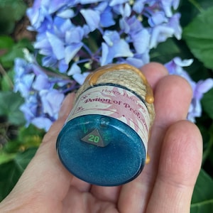 Könnte beinhalten: Ein kleines Glasfläschchen gefüllt mit schimmernder blauer Flüssigkeit, die einen dunkelgrünen zwanzigseitigen Würfel enthält. Der Hals ist mit beigefarbenem Garn und goldenen Wachstropfen umwickelt. Ein Etikett trägt die Aufschrift Happy Hatchling Potion of Probability.